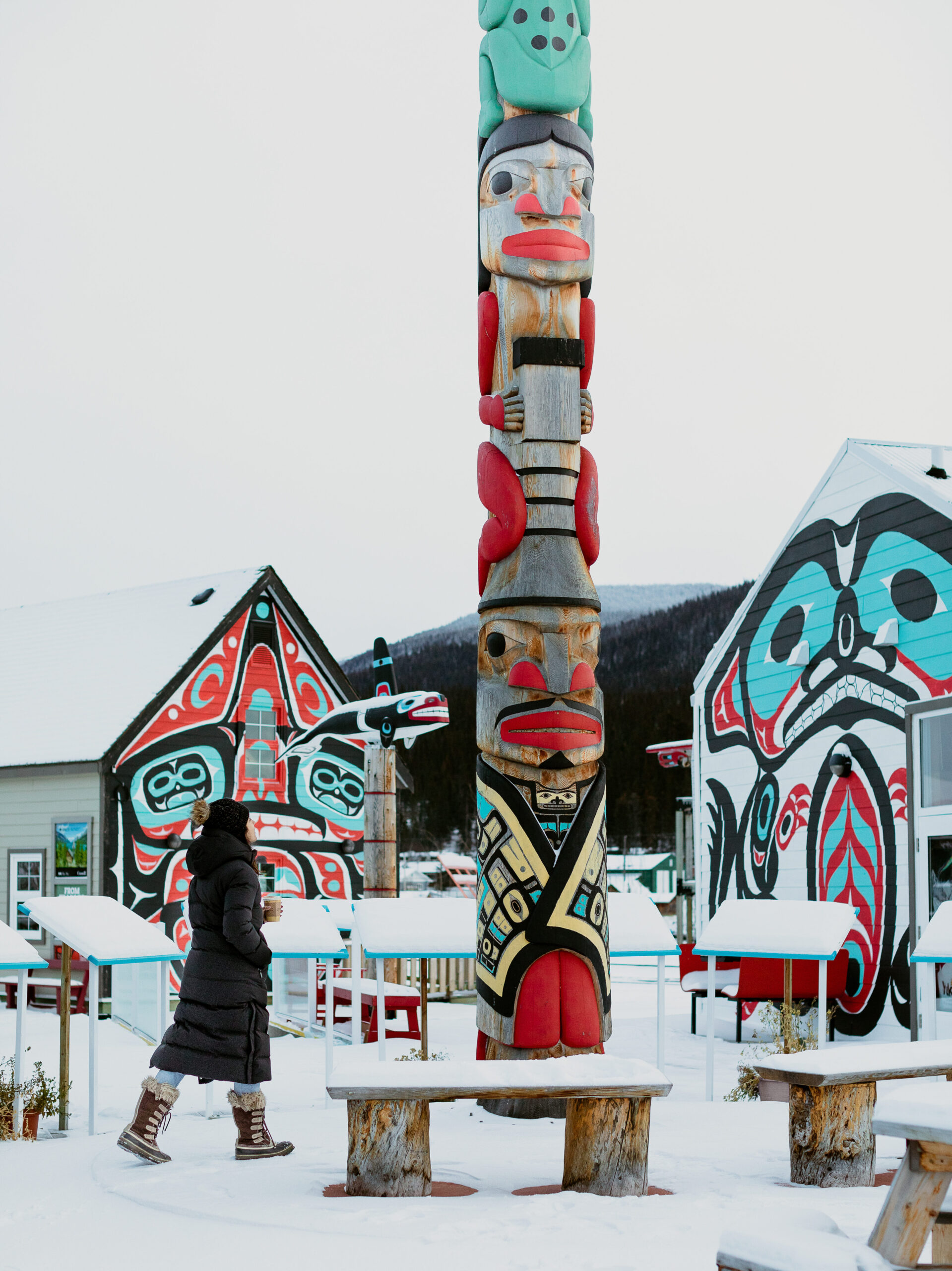 Winter activities in the Yukon. A woman walks through a snowy village - Carcross Commons - with totem pole and other First Nations paintings in the background.