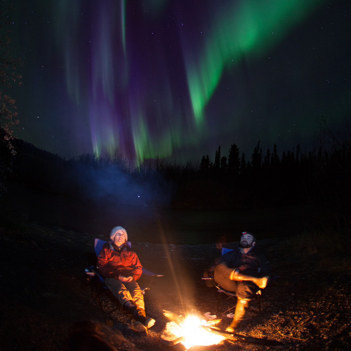 Two people sit around the campfire catching the Aurora/Northern Lights in the Yukon Territory.