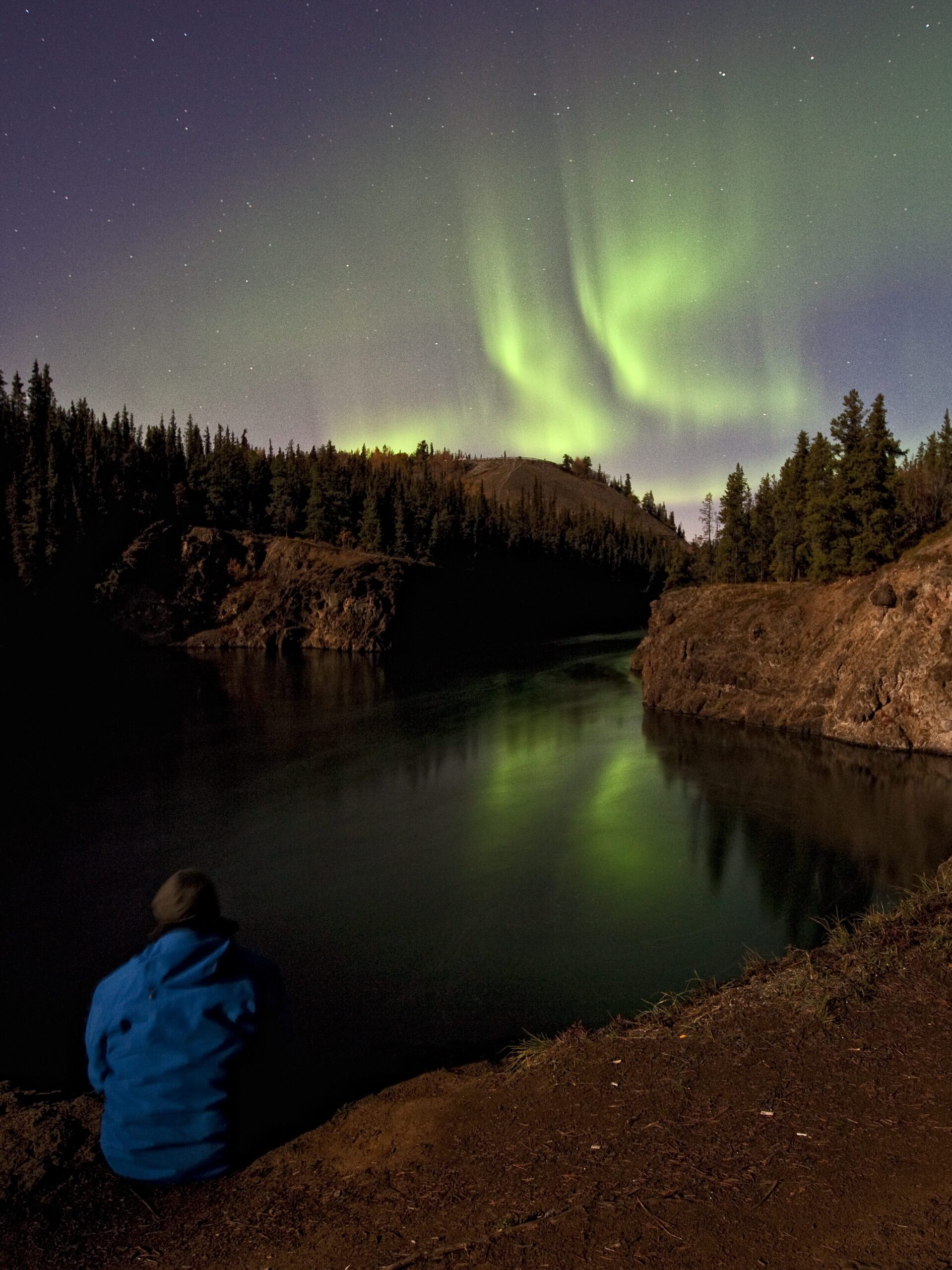 Aurora/Northern Lights from Miles Canyon, Yukon Territory, a person sits on the cliff edge watching.